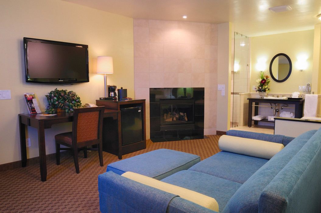A hotel room with a blue sofa, fireplace, desk, TV, and mini-fridge.